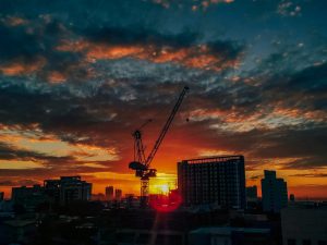 Silhouette of a construction crane and city buildings against a sunset sky with vivid colors, creating a dramatic urban scene.