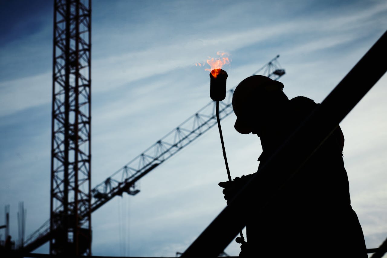 our-services-1 Silhouette of a construction worker using a blowtorch at a building site against a crane-filled skyline.